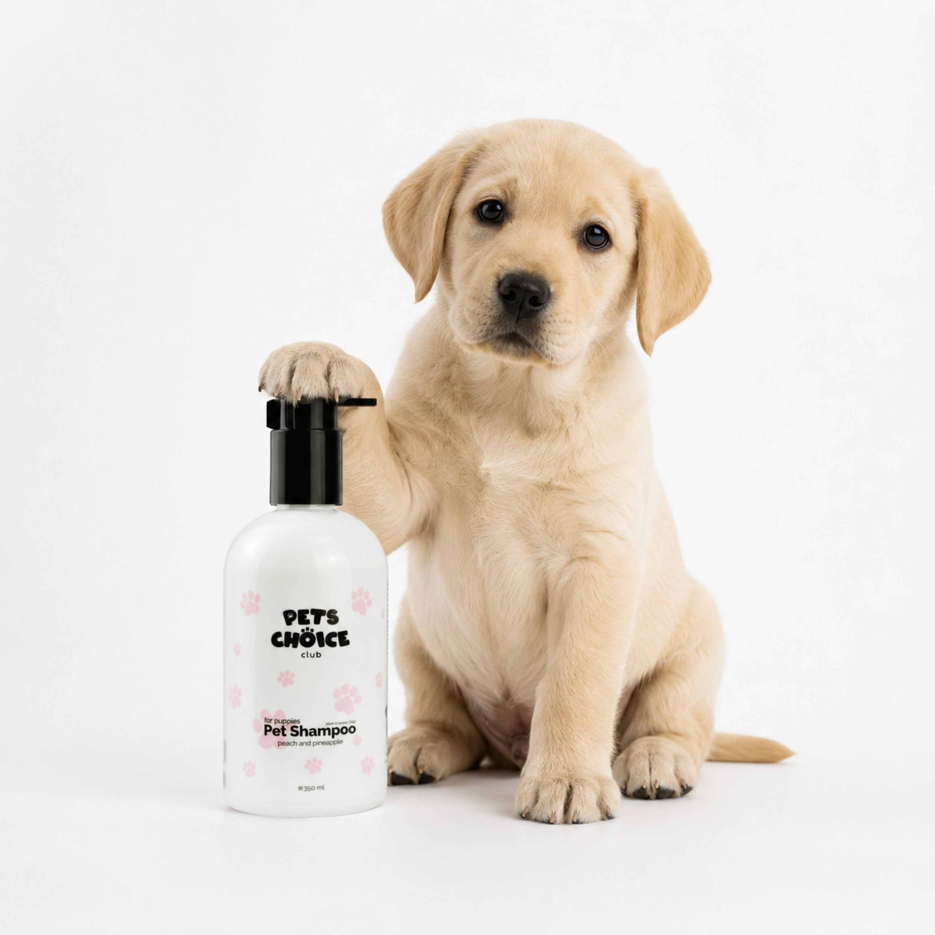 A light cream-colored Labrador puppy sits on a white studio background, resting its right paw on a white bottle of 'Pet's Choice' pet shampoo with a black pump.