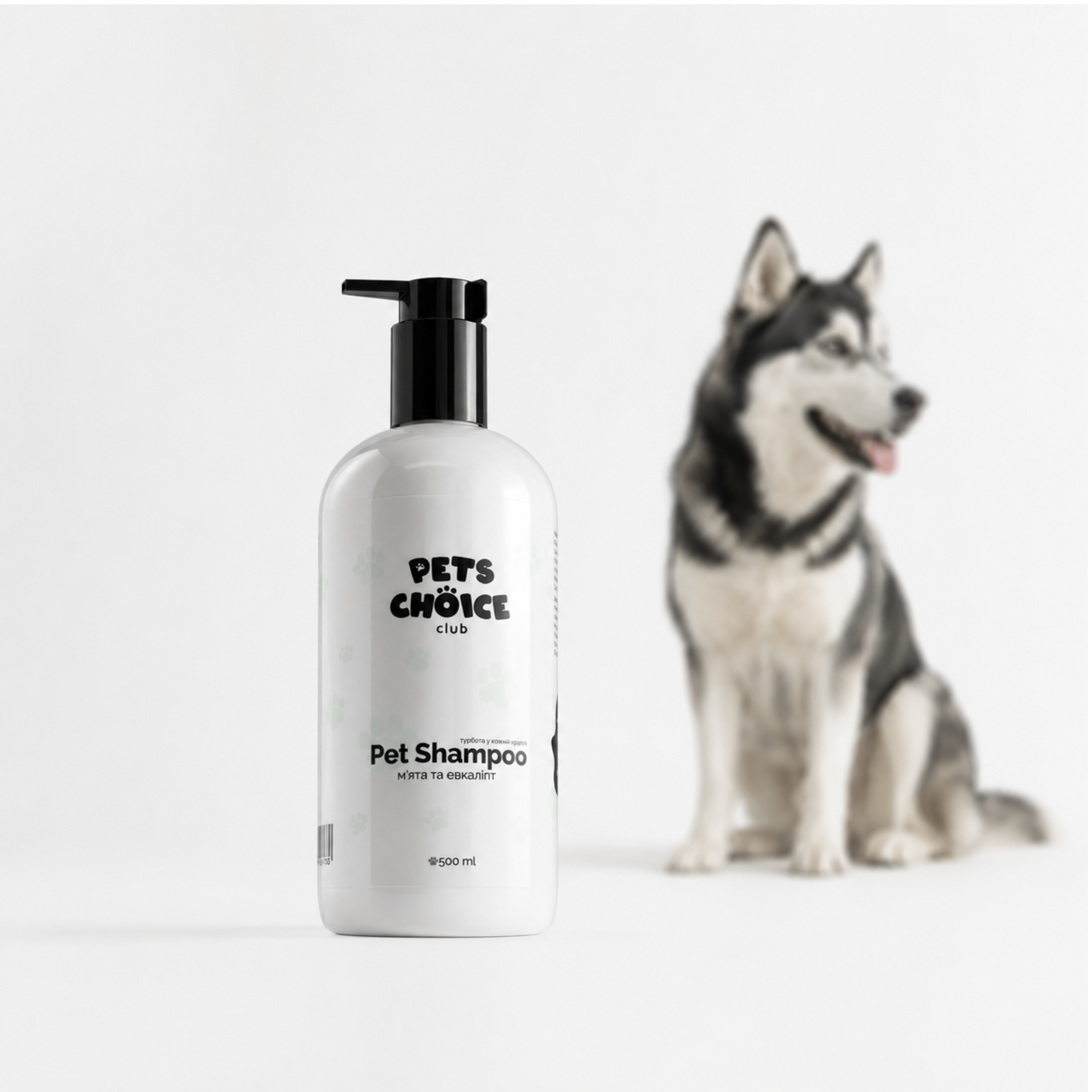 A white bottle of Pet Shampoo with a black pump dispenser, labeled 'PETS CHOICE CLUB', shown in the foreground with a blurred husky dog sitting in the background against a clean white studio backdrop.