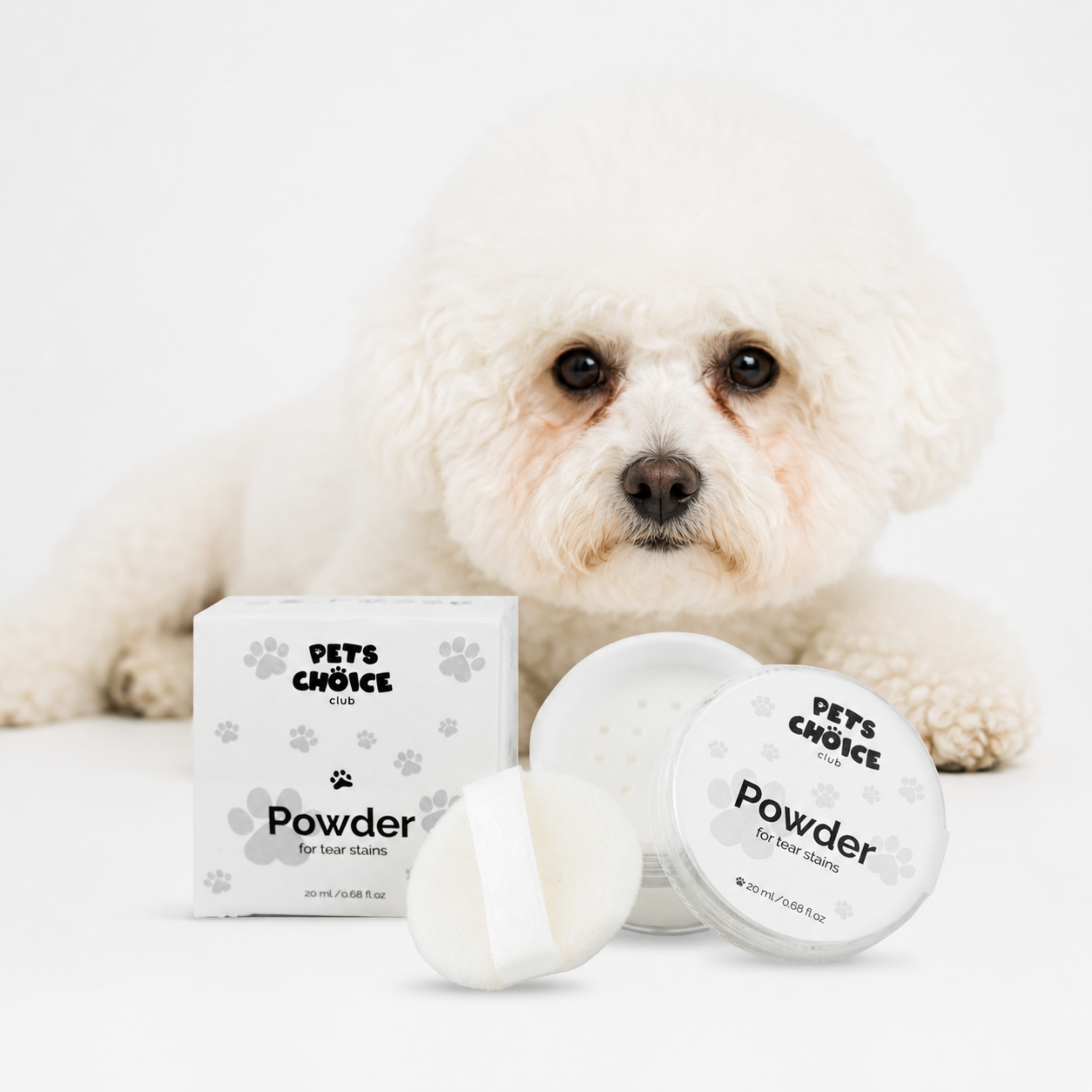 A small white dog lies behind a box and two round jars of Pets Choice Powder pet hair remover on a clean white background.