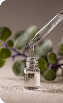 Glass dropper with clear liquid over a small bottle, surrounded by green leaves.